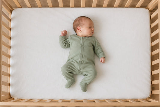 A newborn in a sage sleepsuit lies on their back on a flat mattress in an empty cot. An illustrative photograph generated using AI.